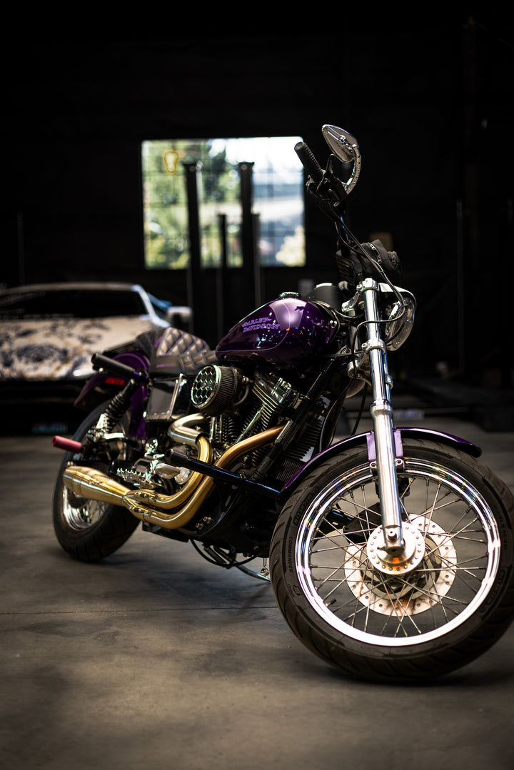 A purple Harley-Davidson Dyna motorcycle with Sawicki Speed 2-into-1 Shorty exhaust in brushed stainless steel finish, parked in a garage with a car int he background.