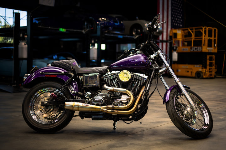 A purple Harley-Davidson Dyna motorcycle with Sawicki Speed 2-into-1 Shorty exhaust in brushed stainless steel finish, parked in a garage with cars and American flag in the background.